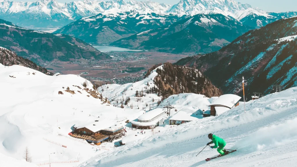 Skijaš u zelenoj jakni spušta se snježnom stazom, a u pozadini su dolina i snježni alpski vrhovi